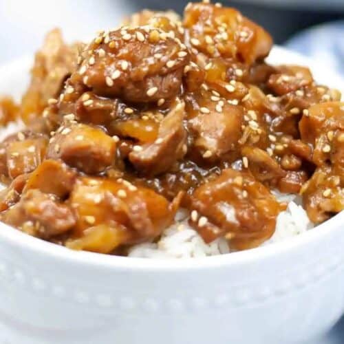 A white bowl filled with white rice topped with pieces of saucy chicken and sprinkled with sesame seeds—this is a must try for anyone who loves Asian dishes.