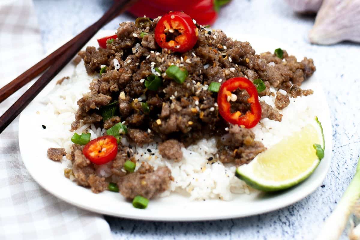 A plate of white rice topped with ground beef, sliced red chili, green onions, and sesame seeds, served with a lime wedge. This must-try dish is better than takeout—chopsticks rest beside the plate for the full Asian dishes experience.