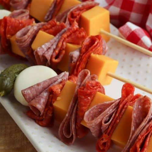 Two charcuterie skewers featuring cubes of cheddar cheese, folded salami, a hard-boiled egg, and a small pickle on a white serving platter.