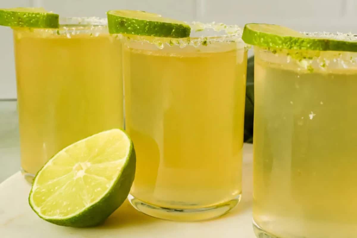 Three glasses of light yellowish-green Jameson shots with salted rims and lime slices sit next to a halved lime on a white surface.