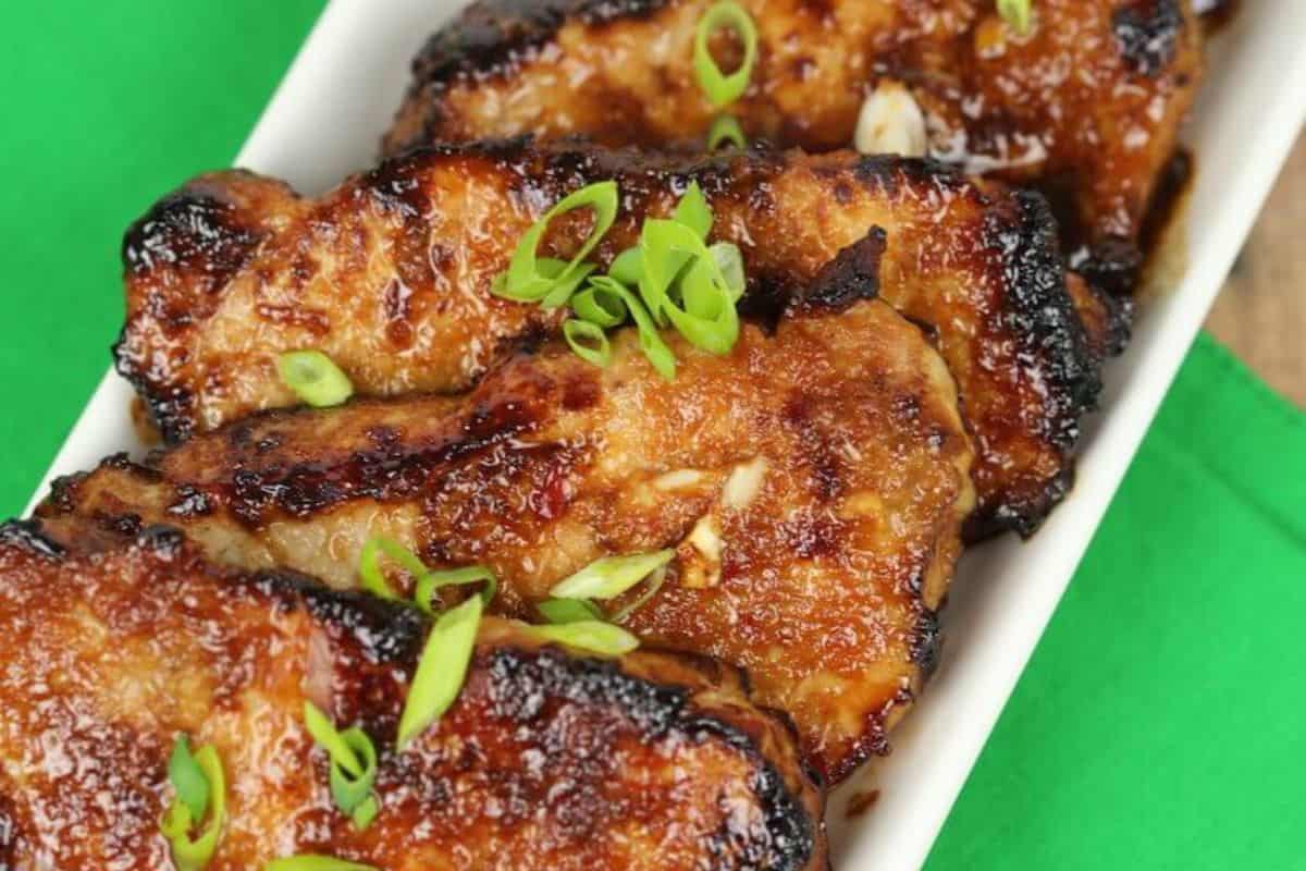 Four glazed pork chops, garnished with sliced green onions, are arranged in a white dish on a green surface—a must try for anyone who loves Asian dishes.