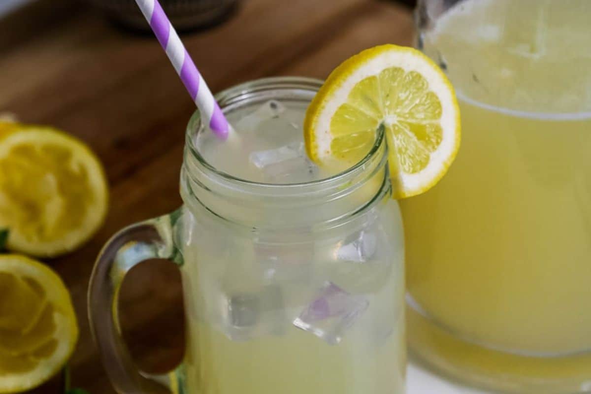 A glass mug filled with lemonade, ice cubes, and a lemon slice sits invitingly. A purple and white striped straw is inserted. In the background, a pitcher hints at secret recipes and halved lemons inspire you to make at home this fresh delight.
