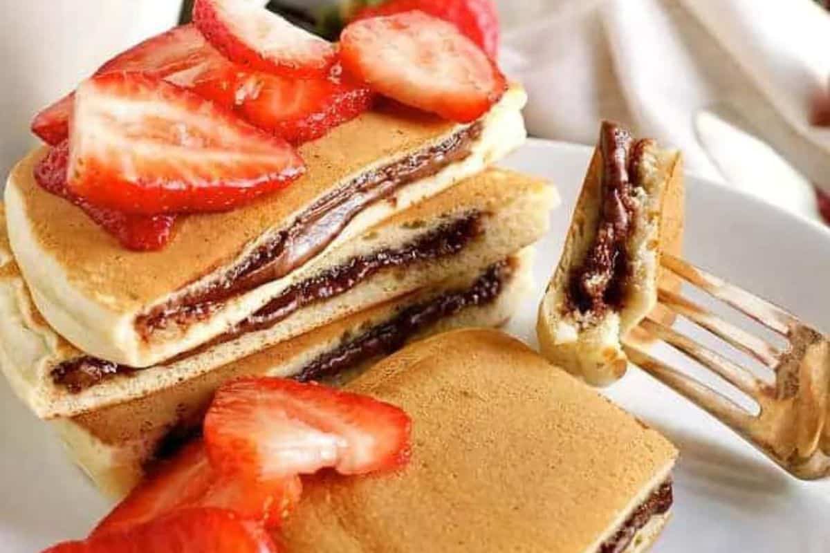 Stack of pancakes filled with chocolate spread, topped with sliced strawberries, on a white plate. A fork holds a piece of the pancake—perfect for those who make every weekend special with delightful pancake recipes.