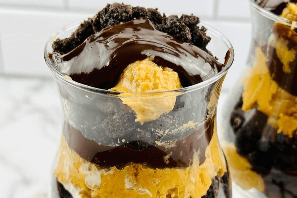 A dessert in a glass with layers of chocolate, crumbled cookies, and a yellow filling, topped with chocolate sauce and cookie crumbles, this treat is one of those must-try recipes.
