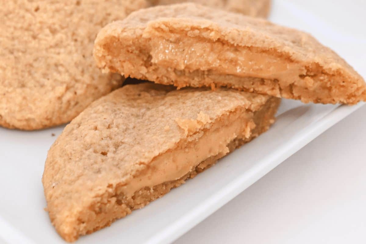 A close-up of peanut butter-filled cookies, one of which is cut in half to reveal a smooth peanut butter center. The cookies, showcasing one of the must-try recipes for any peanut butter enthusiast, are placed on a white plate.