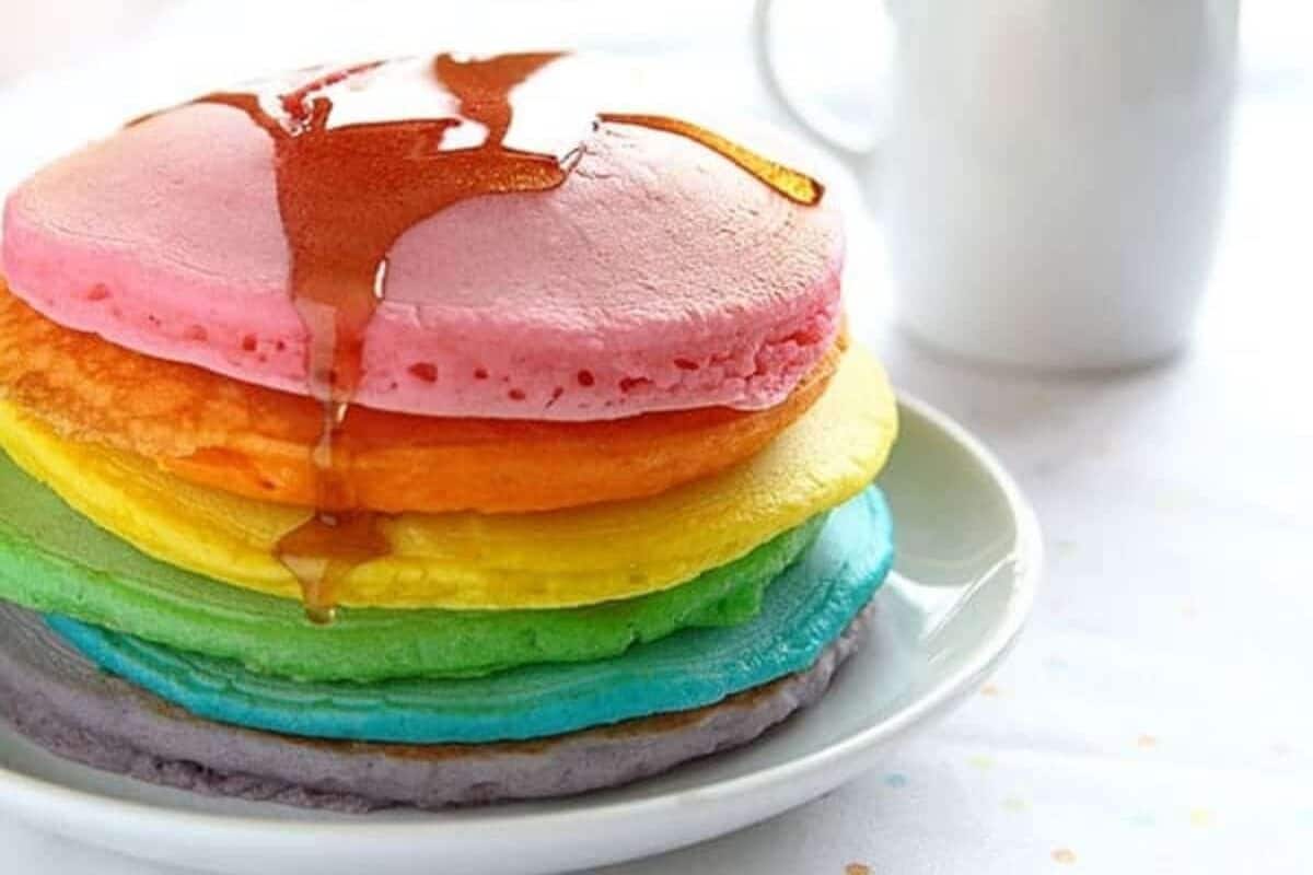 A stack of colorful rainbow pancakes drizzled with syrup sits enticingly on a white plate, perfect for a weekend treat. In the background, a white mug hints at morning bliss, inviting you to make memories with delightful pancake recipes.