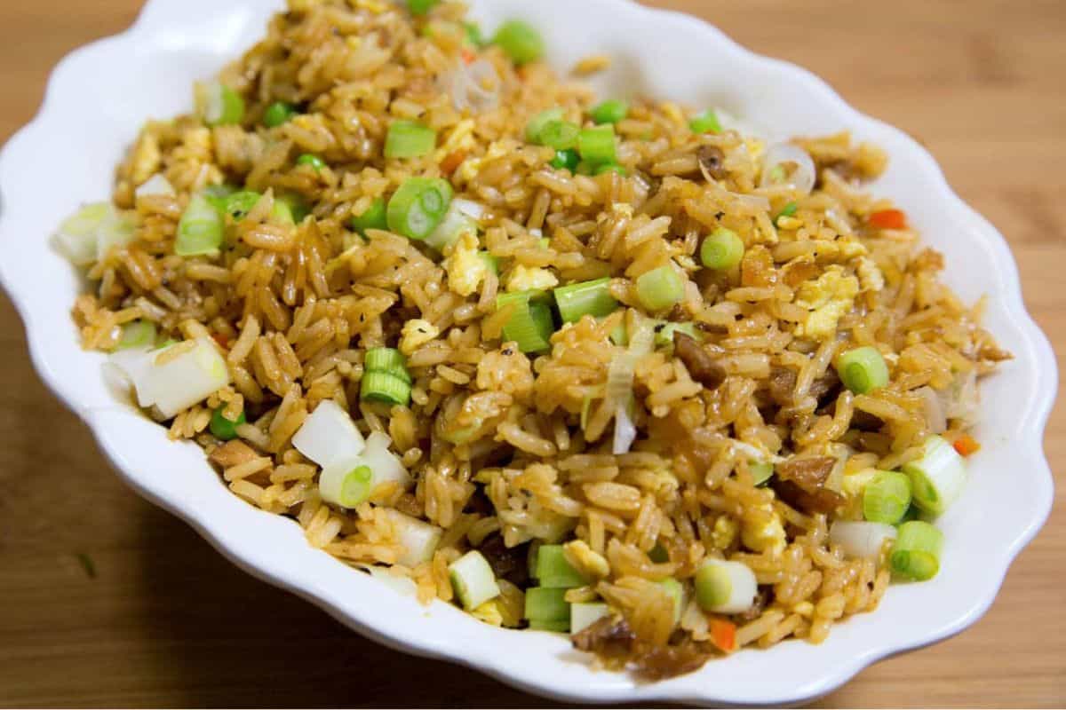 A white dish filled with fried rice, mixed with vegetables and scrambled eggs, and topped with chopped green onions sits on a wooden surface—a must try for fans of Asian dishes.