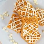 Four crispy Pumpkin Rice Krispie Treats squares drizzled with white icing are arranged on a white plate with scattered white chocolate chips.