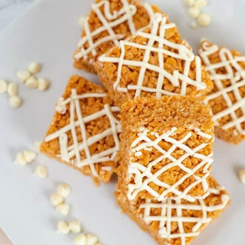 Four crispy Pumpkin Rice Krispie Treats squares drizzled with white icing are arranged on a white plate with scattered white chocolate chips.