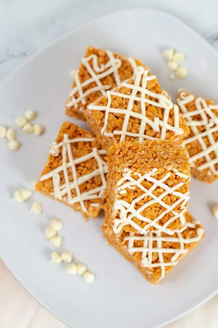 Four crispy Pumpkin Rice Krispie Treats squares drizzled with white icing are arranged on a white plate with scattered white chocolate chips.