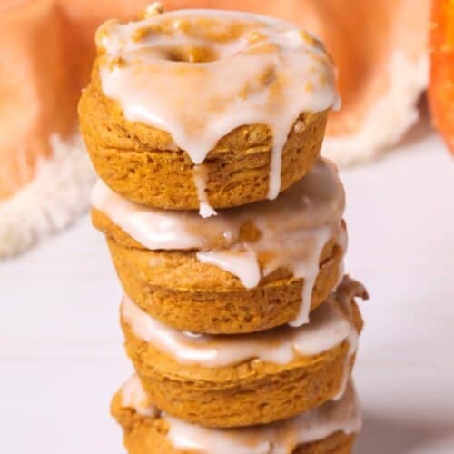 A stack of four pumpkin spice donuts topped with white icing, with an orange cloth and a pumpkin in the background.