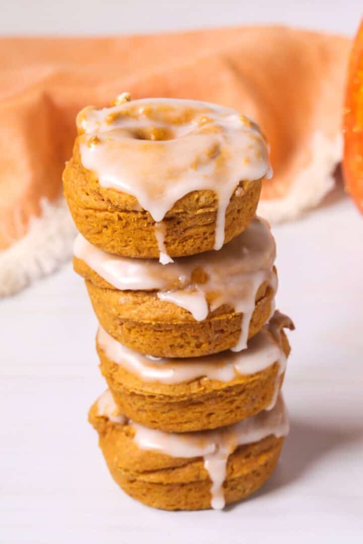 A stack of four pumpkin spice donuts topped with white icing, with an orange cloth and a pumpkin in the background.