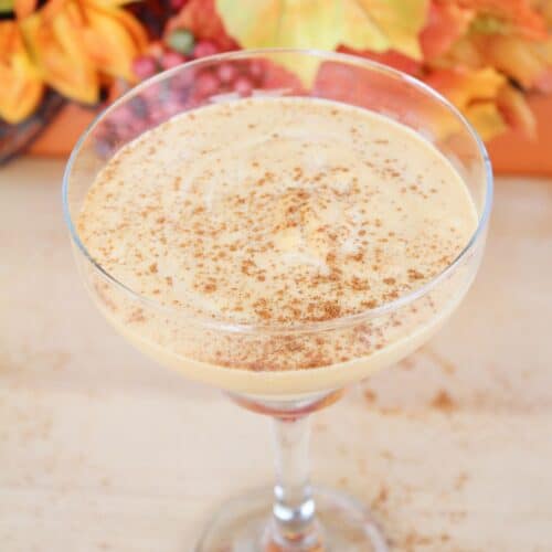 A glass of creamy orange-colored drink, reminiscent of pumpkin spice white russians, is topped with ground spice and surrounded by autumn leaves and orange flowers.