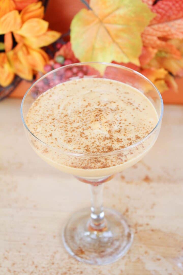 A glass of creamy orange-colored drink, reminiscent of pumpkin spice white russians, is topped with ground spice and surrounded by autumn leaves and orange flowers.