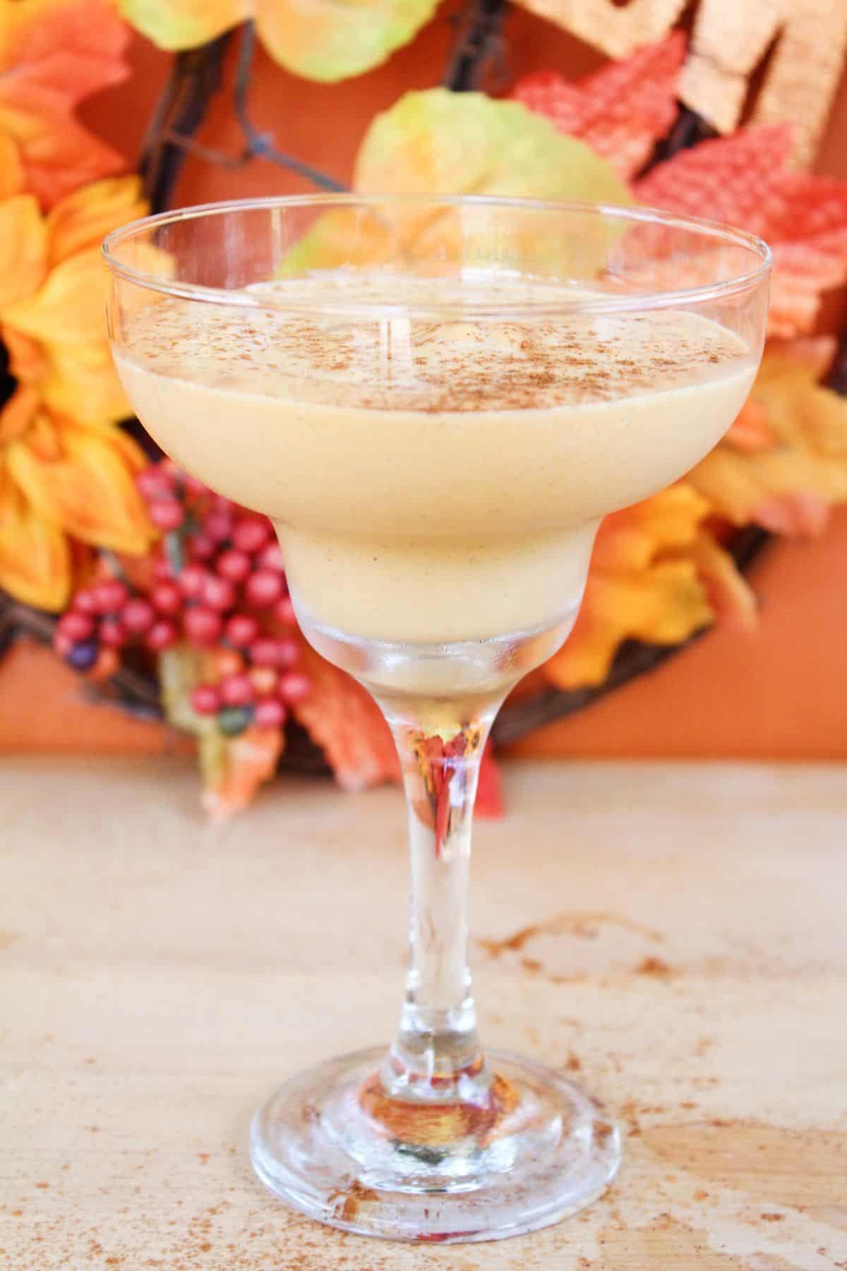 A creamy Pumpkin Spice White Russian cocktail in a margarita glass, topped with ground cinnamon, sits on a light wooden surface with autumn leaves in the background.