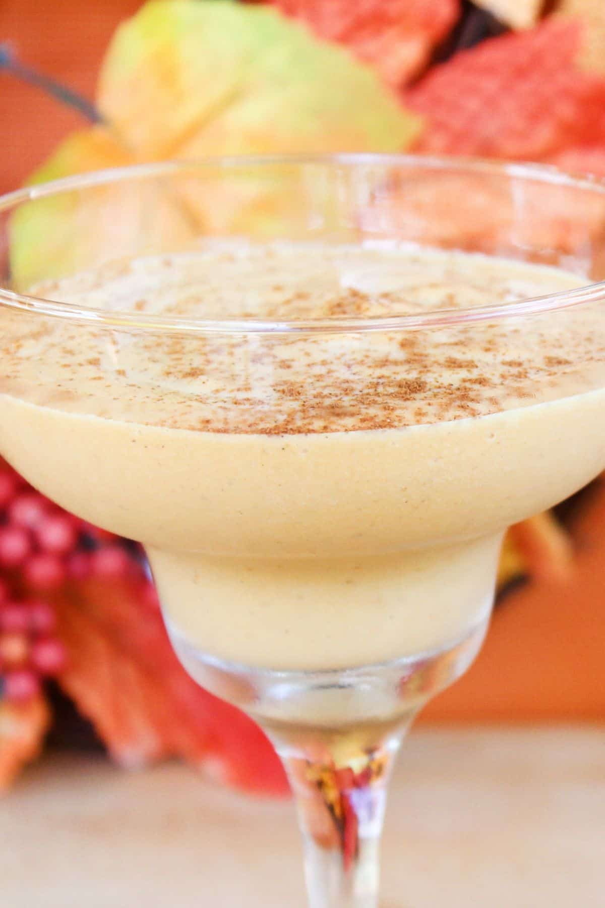 A creamy pumpkin-colored Pumpkin Spice White Russian in a margarita glass, topped with a sprinkle of cinnamon, with autumn leaves in the blurred background.