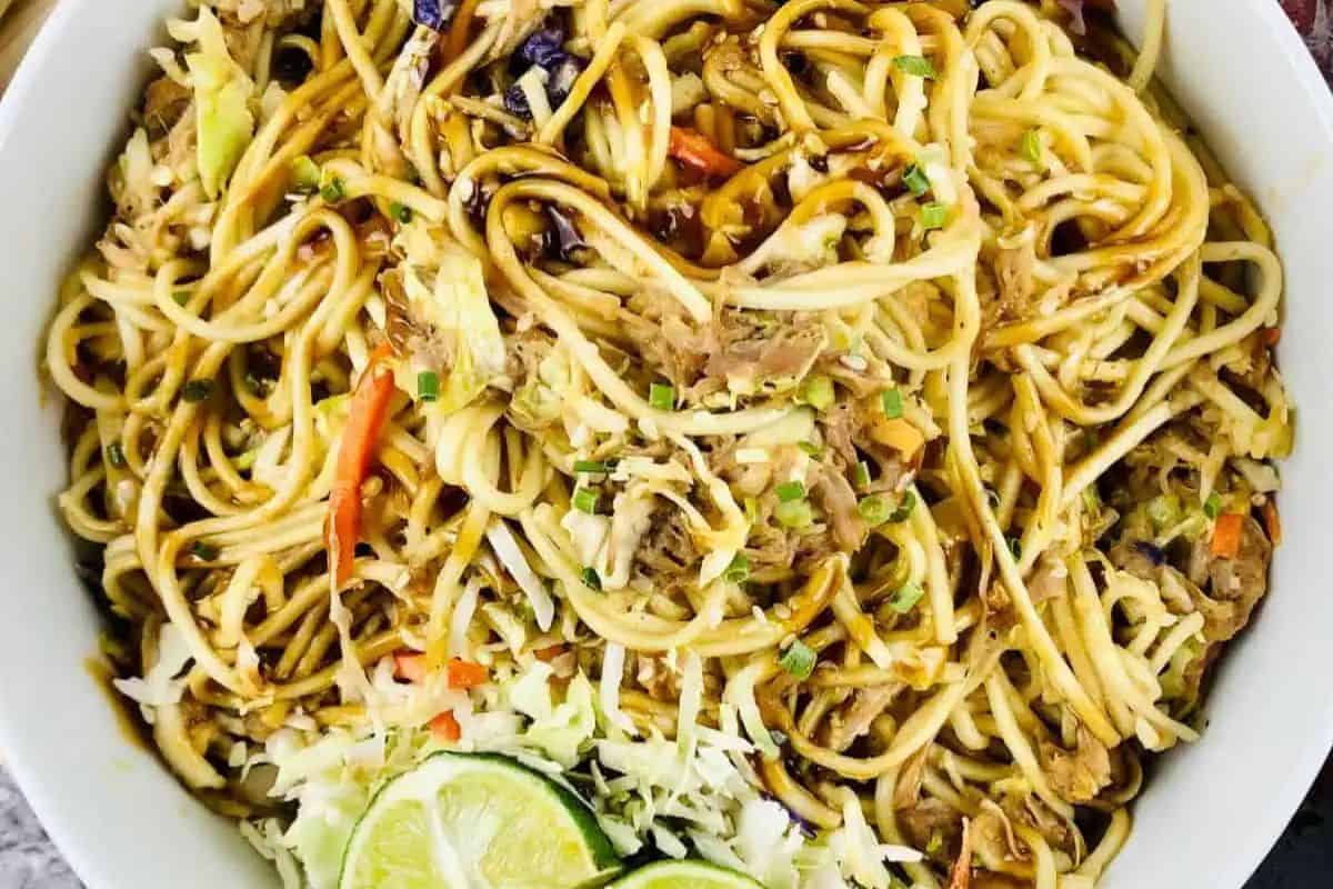A bowl of stir-fried noodles with shredded vegetables, sliced lime, and sauce, garnished with chopped green onions—a must try for fans of Asian dishes.