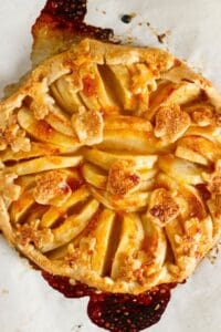 A rustic apple tart with a golden flaky crust, arranged apple slices, and cut-out leaf shapes, rests on parchment paper with caramelized juices. This baked apple galette beautifully combines elegance and homestyle charm.