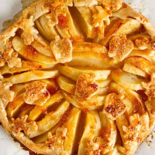A rustic apple tart with a golden flaky crust, arranged apple slices, and cut-out leaf shapes, rests on parchment paper with caramelized juices. This baked apple galette beautifully combines elegance and homestyle charm.