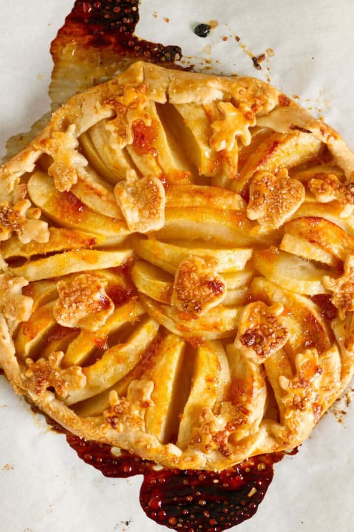 A rustic apple tart with a golden flaky crust, arranged apple slices, and cut-out leaf shapes, rests on parchment paper with caramelized juices. This baked apple galette beautifully combines elegance and homestyle charm.