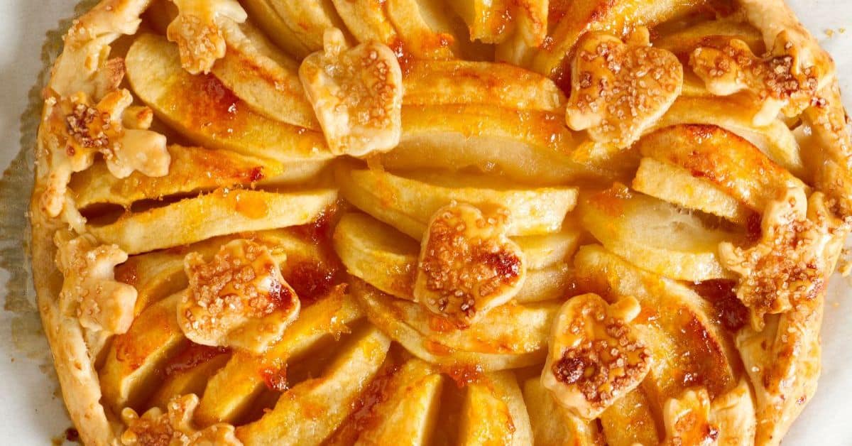 A rustic apple tart featuring golden, overlapping apple slices and small, leaf-shaped pastry pieces on top, all beautifully glazed for a shiny finish.