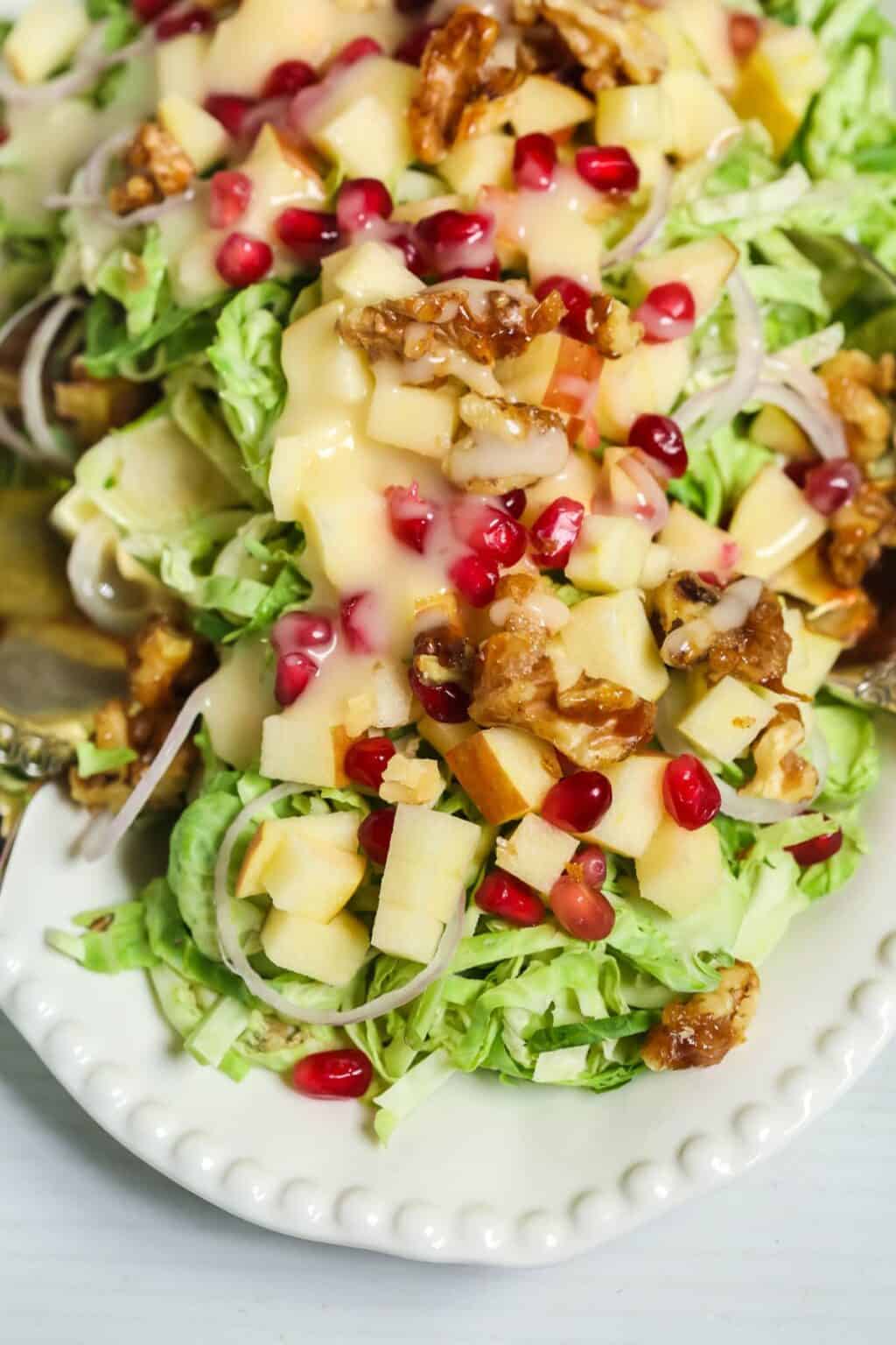 Shaved Brussel Sprouts Salad with Apples features shredded lettuce, crisp chopped apples, pomegranate seeds, candied walnuts, and creamy dressing on a white plate.