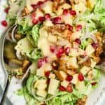 A white plate with Shaved Brussel Sprouts Salad with Apples, walnuts, pomegranate seeds, and sliced onions, all topped with creamy dressing. Two serving spoons rest on the plate.