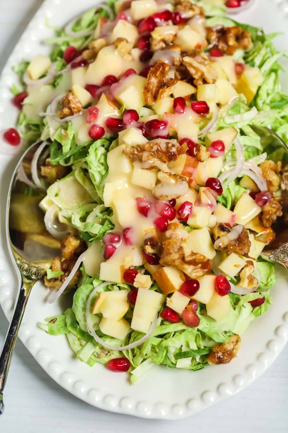 A white plate with Shaved Brussel Sprouts Salad with Apples, walnuts, pomegranate seeds, and sliced onions, all topped with creamy dressing. Two serving spoons rest on the plate.
