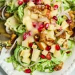 Shaved Brussel Sprouts Salad with Apples features chopped apples, pomegranate seeds, walnuts, shredded greens, and creamy dressing, all served on a white plate.
