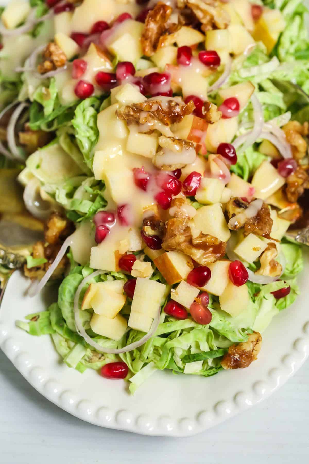 Shaved Brussel Sprouts Salad with Apples features chopped apples, pomegranate seeds, walnuts, shredded greens, and creamy dressing, all served on a white plate.