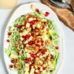 A white oval plate features the dish pomegranate seeds and walnuts. A pitcher of dressing and serving spoons are beside the plate.