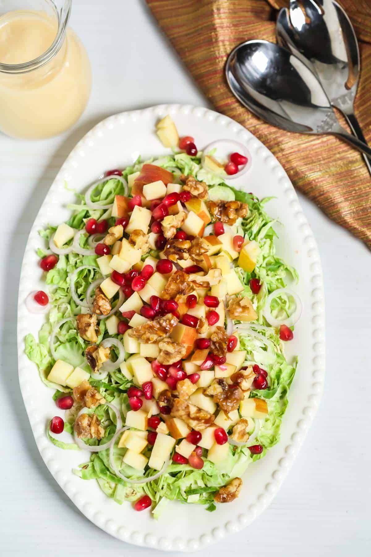 A white oval plate features the dish pomegranate seeds and walnuts. A pitcher of dressing and serving spoons are beside the plate.