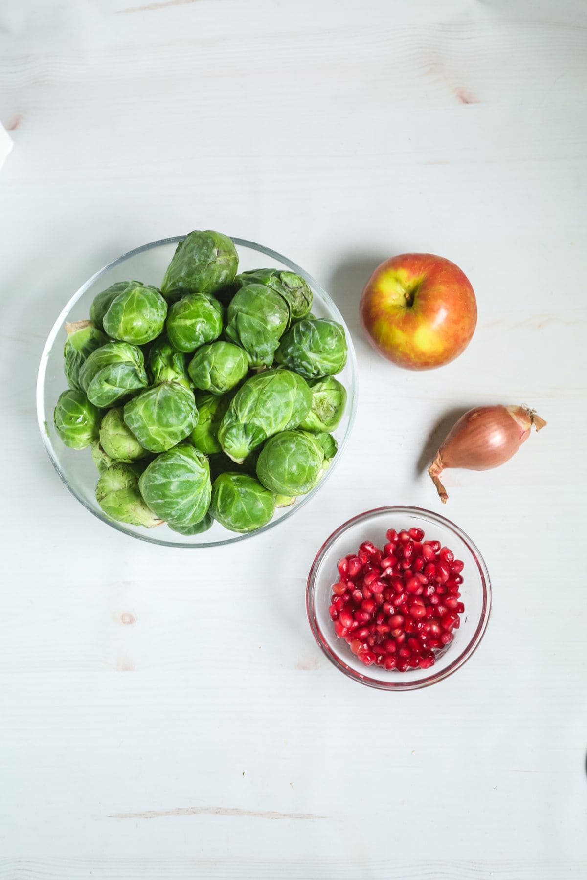 A glass bowl of Shaved Brussel Sprouts Salad with Apples, a glass bowl of pomegranate seeds, a whole apple, and a shallot on a white surface.