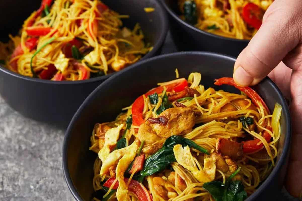 Three black bowls filled with stir-fried noodles, chicken, red bell peppers, and leafy greens are arranged on a gray surface; a hand holds one bowl. These Asian dishes are a must try and taste even better than takeout.