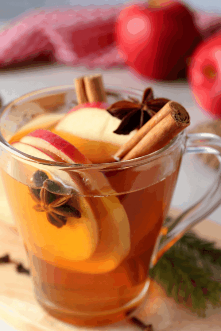 Clear glass mug of slow cooker cider garnished with apple slices, cinnamon sticks, and star anise, with red apples and a red cloth in the background.