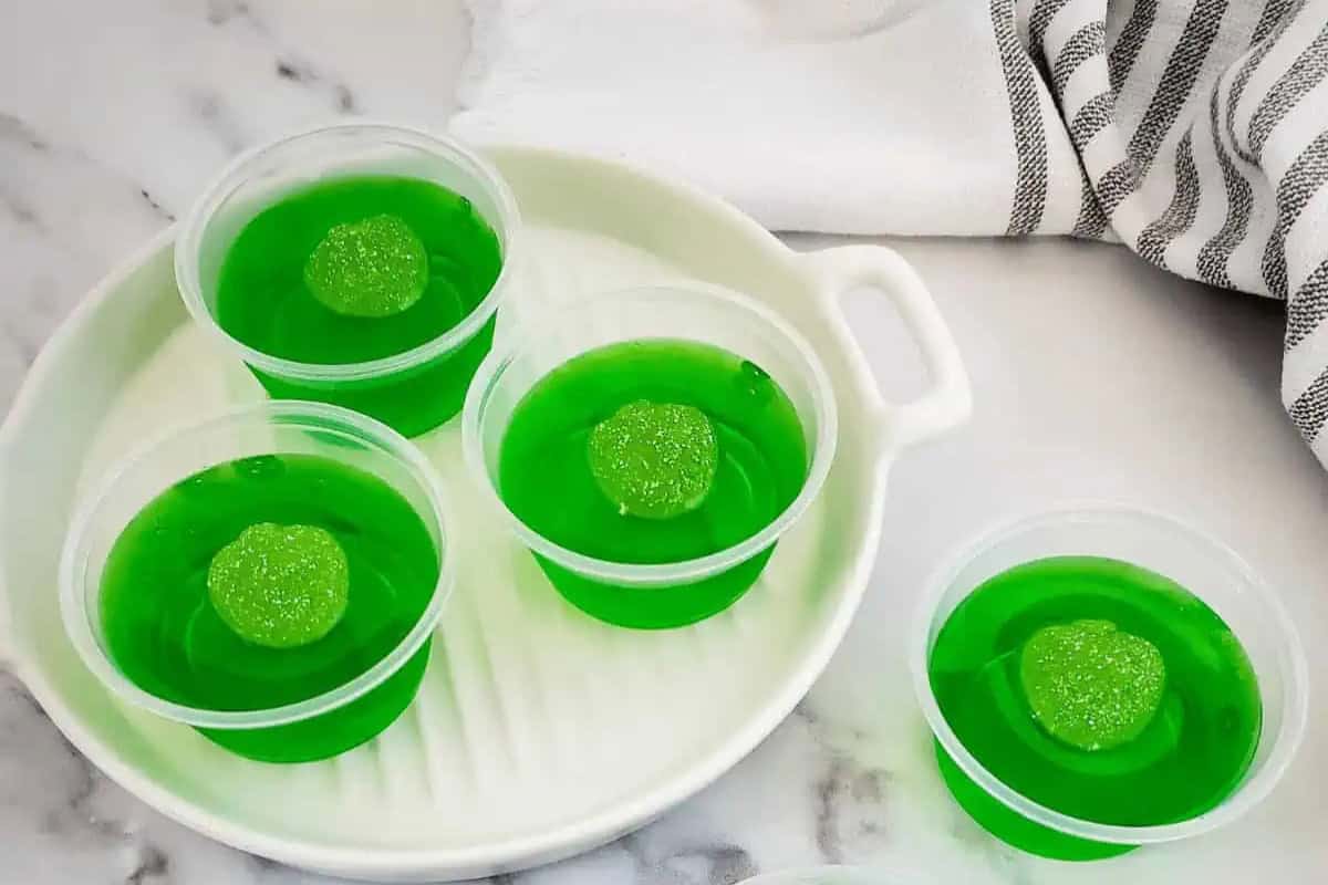 Four small plastic cups filled with green Jello shots, each topped with a green apple gummy candy, arranged on a white tray with a striped towel nearby.