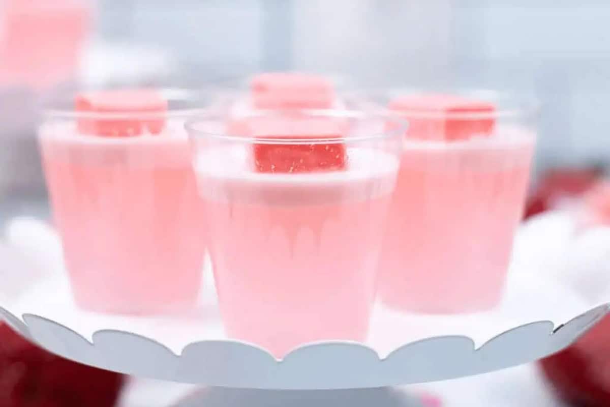 Four clear plastic cups filled with strawberry Jello shots and topped with pink Starburst are displayed on a white scalloped tray.