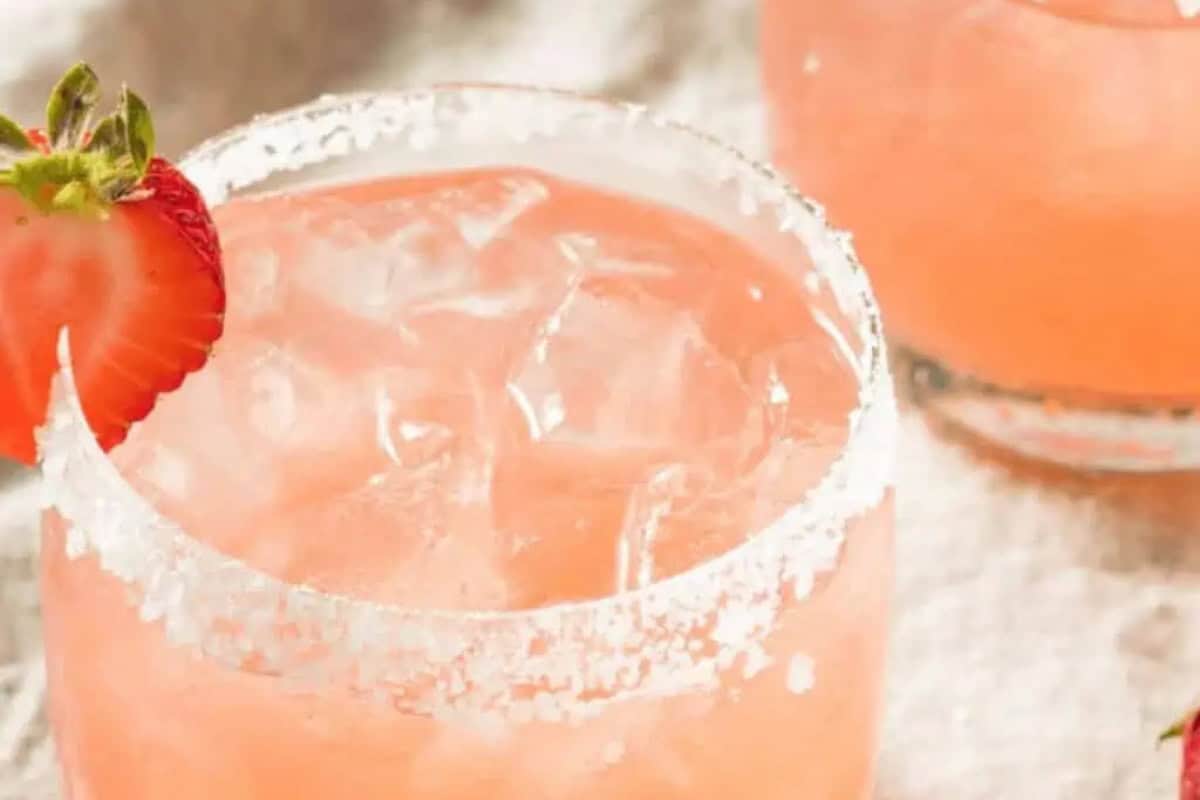 A glass of pink strawberry margarita with ice, a salted rim, and a strawberry garnish on the side. Another similar glass sits in the background, ready to elevate your next gathering.