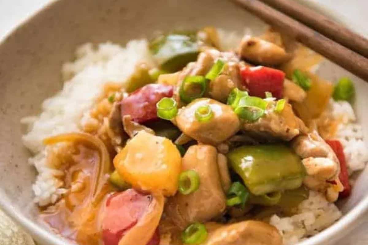 A bowl of white rice topped with stir-fried chicken, bell peppers, pineapple, onions, and sliced green onions—this must try dish is one of those Asian dishes that's better than takeout, with chopsticks resting on the bowl's edge.