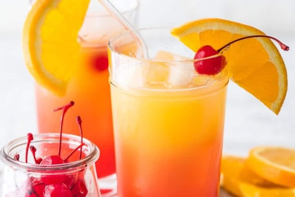 Two glasses of vibrant tequila sunrise Cocktails with orange and red layers, iced and garnished with orange slices and cherries, sit beside a jar of maraschino cherries and fresh orange slices.