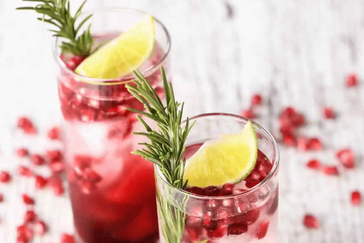 Two glasses filled with a red pomegranate cocktail drink, ice, pomegranate seeds, garnished with lime wedges and rosemary sprigs.