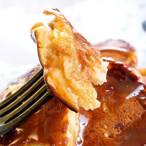 A fork lifts a bite of apple cinnamon pancakes drizzled with syrup from a stack.