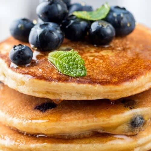 Enjoy a weekend treat with a stack of pancakes topped with blueberries and a mint leaf, drizzled with syrup. Perfect for those exploring new pancake recipes!