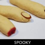 Three witch finger cookies made of dough, with almond slices as fingernails and red jelly or jam at the tips, are arranged on a white surface for a spooky witch finger cookies display.