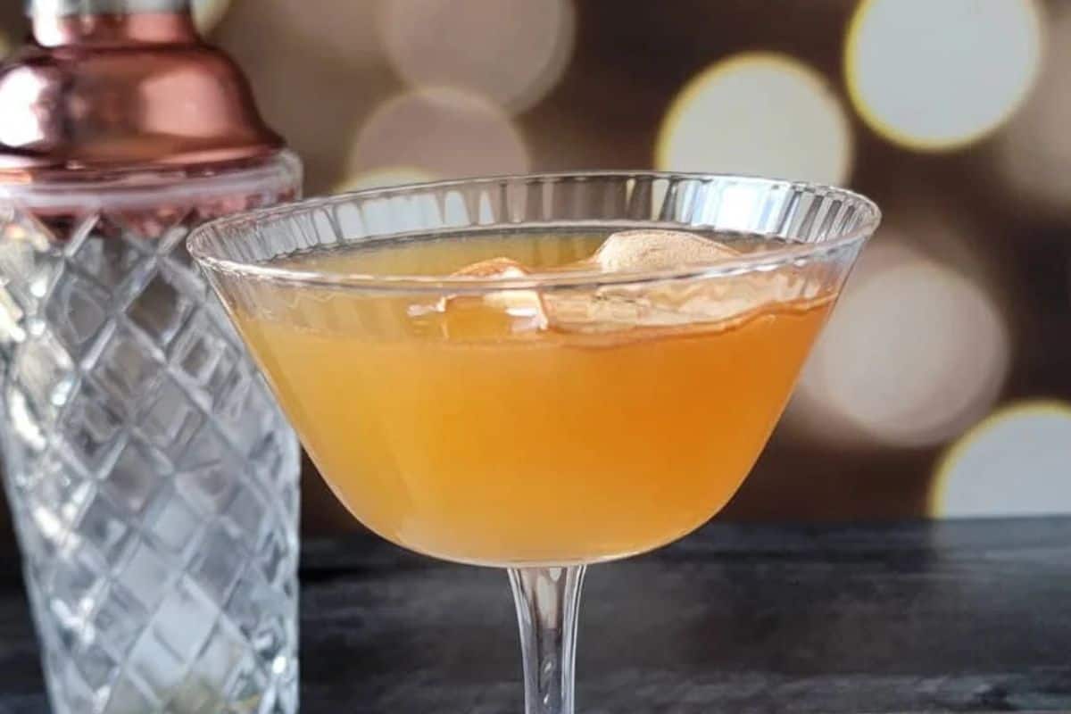 A coupe glass filled with an orange autumn cocktail and ice sits beside a patterned shaker on a dark surface, with blurred lights glowing in the background.