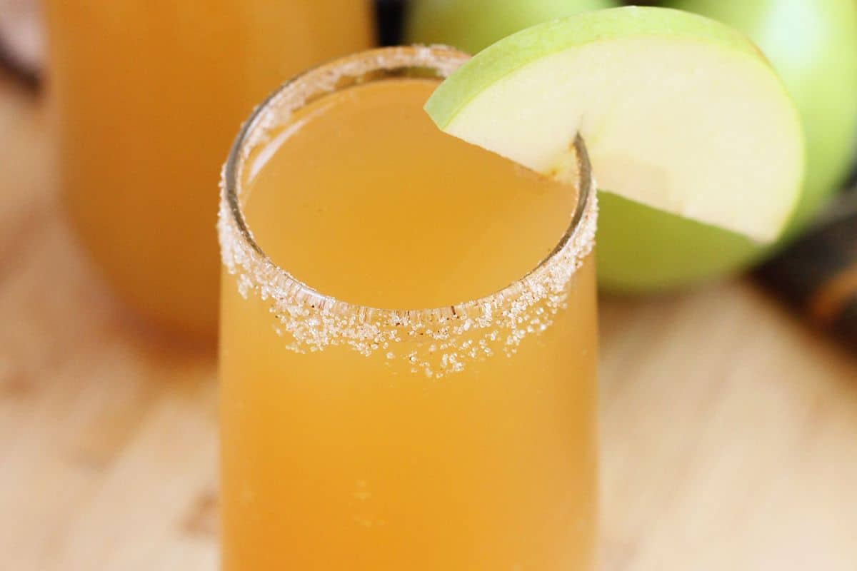A glass of orange-brown seasonal cocktail with a sugared rim, garnished with a slice of green apple, sits on a wooden surface.