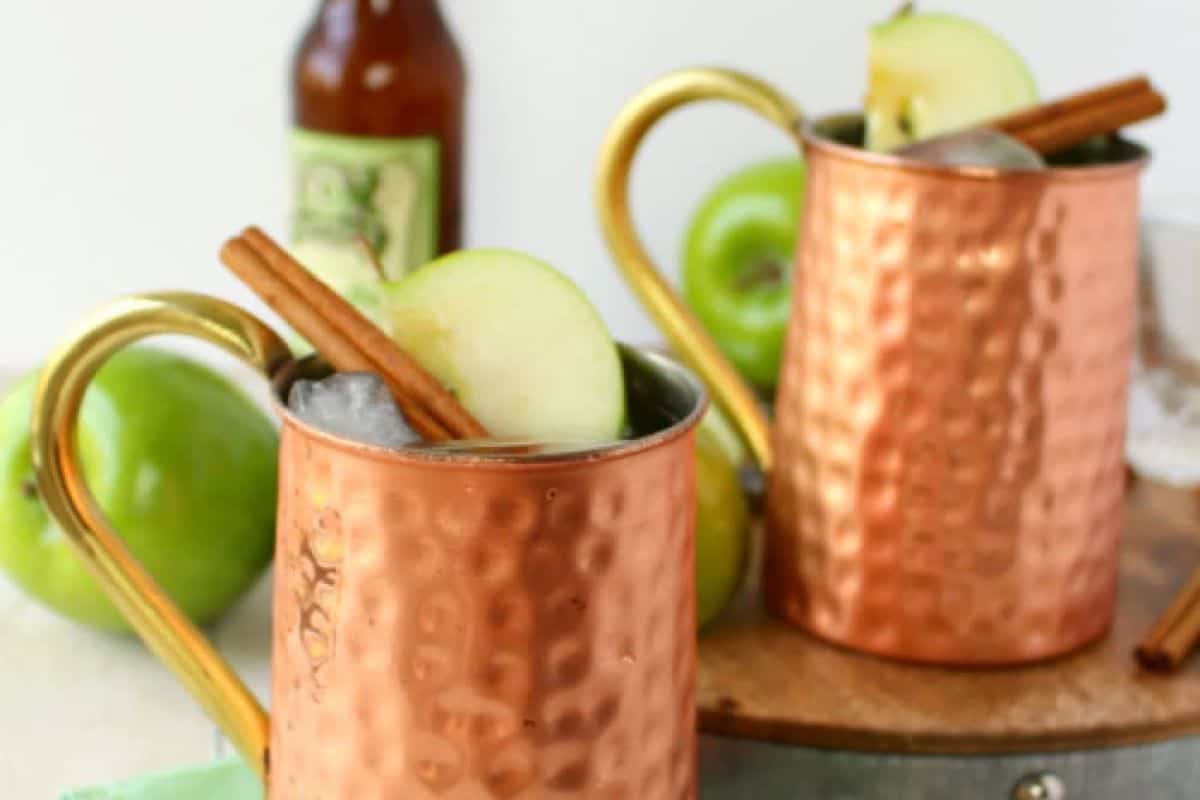 Two copper mugs filled with ice, garnished with green apple slices and cinnamon sticks—perfect for autumn cocktails. A fresh green apple and a bottled drink complete this inviting fall drinks setup.