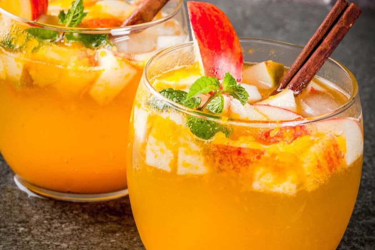 Two glasses of orange-colored autumn cocktails with ice cubes, apple slices, mint leaves, and cinnamon sticks rest on a dark surface.
