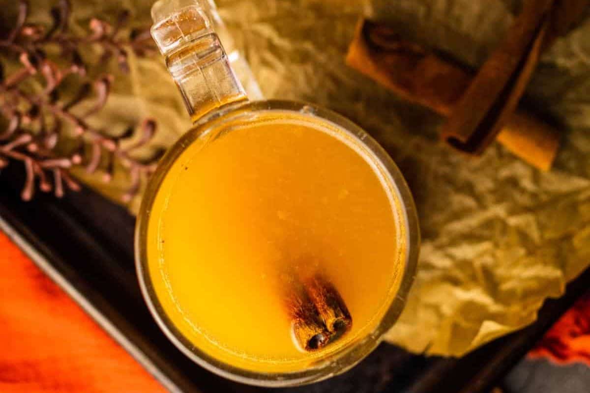 A clear glass mug filled with a light brown beverage, garnished with a cinnamon stick, sits on a parchment-lined surface with extra cinnamon sticks nearby—perfect for savoring cozy autumn cocktails.