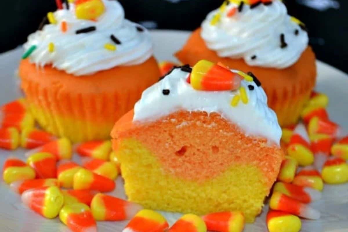  A close-up of a cupcake cut in half, revealing a yellow and orange striped interior. It's topped with white frosting, a candy corn, and colorful sprinkles. Other cupcakes are visible in the background, all surrounded by loose candy corn.